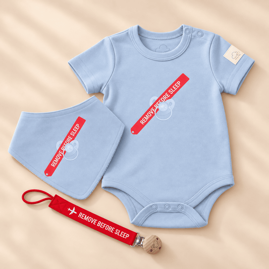A baby clothing set laid flat on a beige fabric background, including a light blue short-sleeve bodysuit, a matching light blue bib, and a red clip with white text reading 'REMOVE BEFORE SLEEP'.