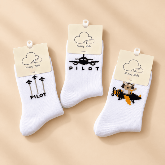 Three pairs of white crew socks with cream paper tags from Kurry Kids, displayed on a warm beige background. The socks feature black and orange graphics: one with 'PILOT' and three arrows, one with a black airplane, and one with a pixel-art bee pilot.